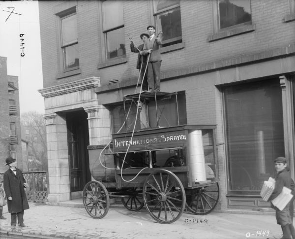 An International Harvester "sprayer" mounted on a wagon, possibly intended for pesticide application (or in this context, window washing?). The wagon is parked on the sidewalk in front of a brick building. The top side of the wagon reads: "International Sprayer". (These words are scratched into the glass plate original rather than painted on the wagon.) Two men are standing on top of the wagon holding the nozzles of the hoses. To the right standing on the sidewalk are two newspaper boys, one of them holding newspapers. On the left a man is looking up. There is snow on the ground and everyone is wearing winter clothing and coats. This may have been taken in Auburn, New York.