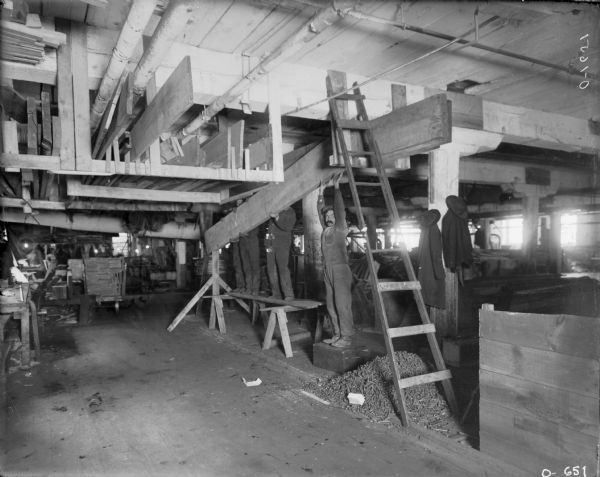 Four men are lifting a large piece of beam during the construction of a factory building, most likely at International Harvester's Osborne Works. There is a ladder leaning against the ceiling rafters, various construction materials are around the room, and lumber is stored in a loft. The men are all wearing work clothes and their hats and coats are hanging on a beam.