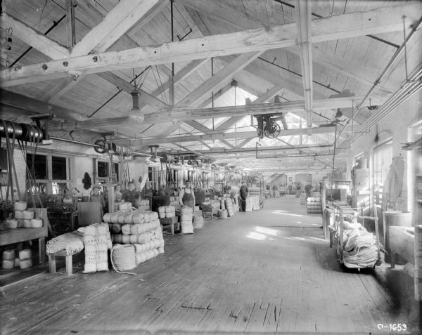 View down the center aisle of International Harvester's Osborne Twine Mill. On either side of the aisle factory workers are producing balls of twine. The factory floor is open and bright with many windows, and rafters hanging from the ceiling. The men and women are wearing work clothes.