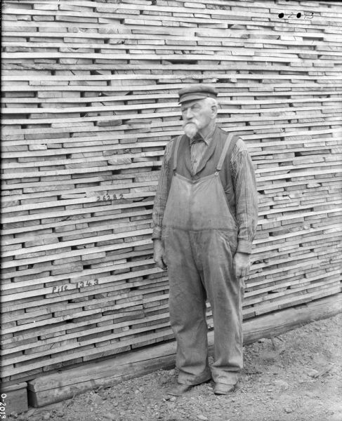 An elderly factory worker is standing in front of large stacked pile of wood at International Harvester's Osborne Works. There are two handwritten notes on the ends of the wood, the lower reading: "Pile 1243" and the upper reading: "2682". The man is wearing overalls, a vest, a button-up shirt, a dark hat, and has a white beard and moustache.