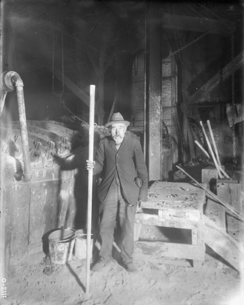 An elderly factory worker is standing in a dusty room holding a large pole at International Harvester's Osborne Works. He is dressed in dark work pants, a dark jacket, and a dark hat. Behind him are buckets, a wooden table, pipes and chains.