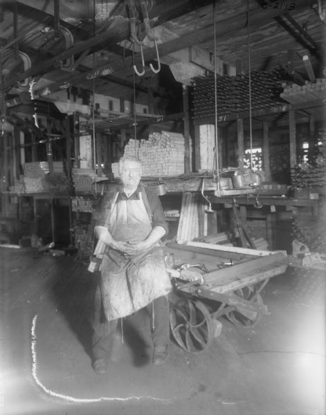 A factory worker is sitting on a cart on at International Harvester's Osborne Works. Behind him are stacked pieces of lumber. Hooks hang from the ceiling. He is wearing dark work clothes, an apron, and a chain around his neck.