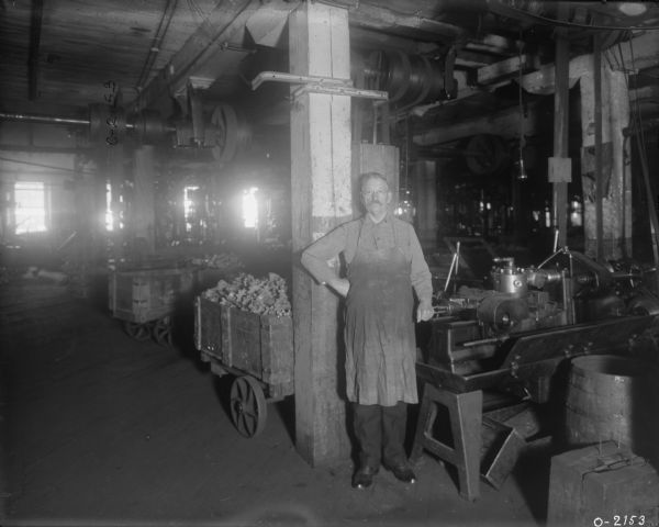 A factory worker is standing next to a large piece of machinery at International Harvester's Osborne Works (later known as Auburn Works). Around him are carts full of parts, and barrels. Belt-driven machinery is attached to the ceiling. The man is dressed in dark work clothes, an apron, and has a moustache.