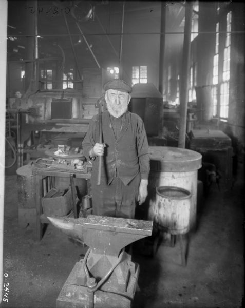 A factory worker with a white beard stands behind an anvil with a large hammer over his shoulder at International Harvester's Osborne Works (later known as Auburn Works). He is dressed in dark clothing: pants, a button-up shirt, vest and hat. In the background are various pieces of manufacturing machinery.