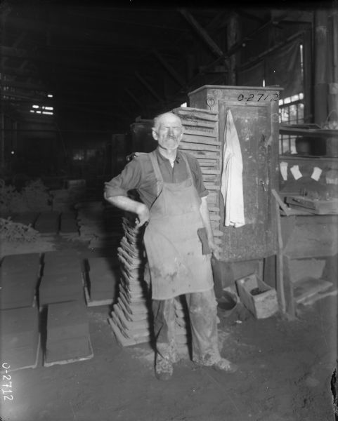 A factory worker wearing work clothes and an apron stands with his arm on his hip at International Harvester's Osborne Works (later known as Auburn Works). Behind him are stacked building materials and pallets. There is a cabinet and desk beside him with a white coat slung over the cabinet.