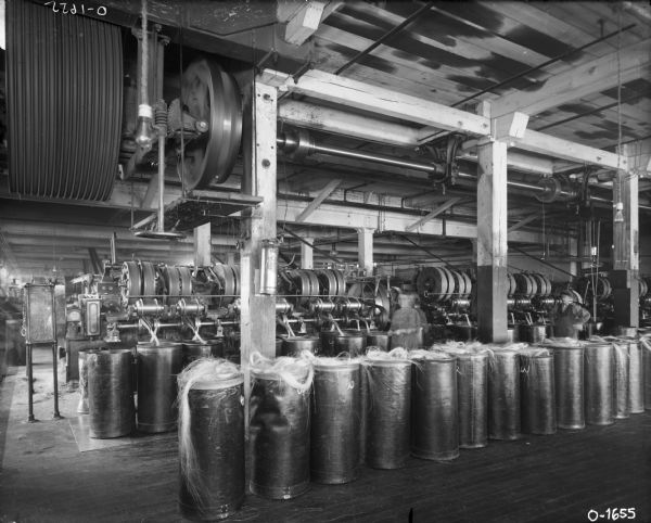 Factory workers, possibly including two women, are wearing work clothes while standing at work stations in a room filled with large machinery at International Harvester's Osborne Works (later known as Auburn Works). In the foreground are tall canisters filled with what appears to be fiber for making twine.