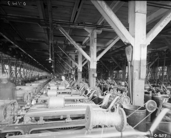 Male and female factory workers are standing at work stations next to large pieces of manufacturing equipment at International Harvester's Osborne Works (later known as Auburn Works). Spools of twine are on the top of the machines. The room is large and filled with machinery, and there are electric lights hanging from the ceiling.