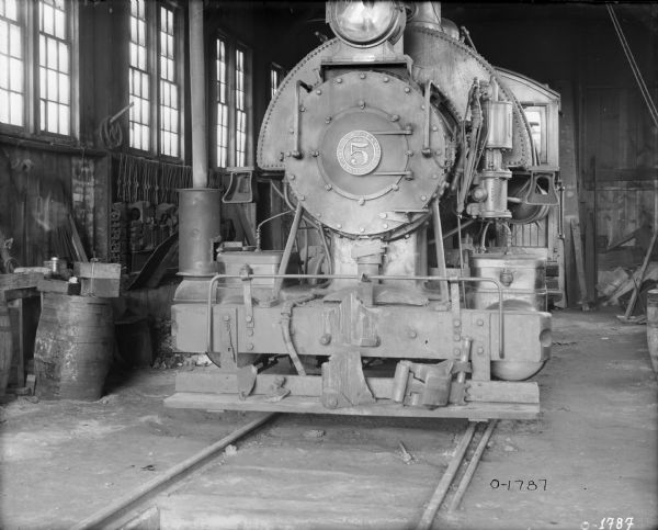 A locomotive is backed into a loading area at International Harvester's Osborne Works (later known as Auburn Works). The locomotive has a large light on the front and the number "5". In the loading area are barrels and chains and tools hanging from the walls.