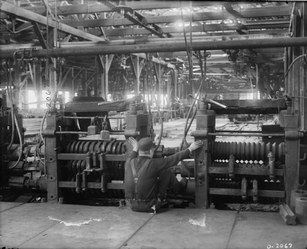 A factory worker is sitting on the ground, bracing himself against large machinery with his feet and hands, at International Harvester's Osborne Works (later known as Auburn Works). He is wearing dark work clothes, suspenders and a hat. The room is large and filled with several different pieces of machinery.