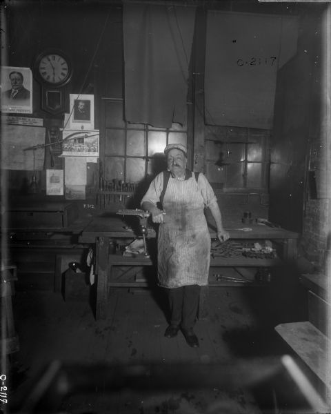 A man wearing work clothes, a hat, and a long apron is standing and posing in front of a work station at International Harvester's Osborne Works (later known as Auburn Works). The table behind him has various tools on it. The windows are covered in makeshift curtains. In the background on the left is a poster of President Taft reading: "Our Next President," as well as calendars, other posters and a clock.