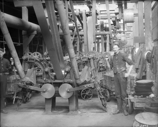 Two factory workers in dark work clothes and hats stand near large, belt-driven manufacturing equipment at International Harvester's Osborne Works (later known as Auburn Works). On the far right coats and hats are hanging along the wall.