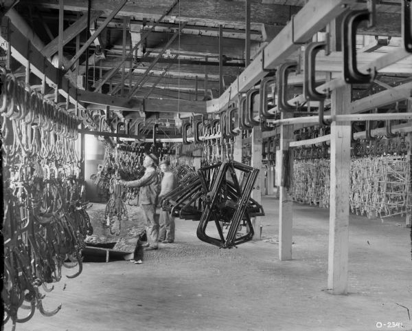 Factory workers are standing among implement parts hanging from hooks suspended from a track on the ceiling at International Harvester's Osborne Works (later known as Auburn Works).