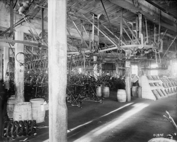 Agricultural machinery parts hang from hooks on a track suspended from a ceiling and winding around a factory floor at International Harvester's Osborne Works (later known as Auburn Works). To the right are slanted wooden storage bins. Barrels located around wooden beams.