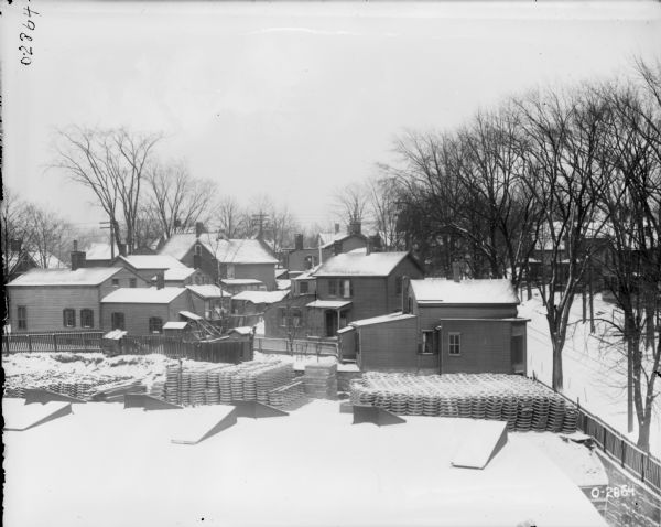 Elevated view of a factory yard with two large piles of stacked implement parts at International Harvester's Osborne Works (later known as Auburn Works). In the background is a residential neighborhood of wooden homes. The ground is covered in snow.