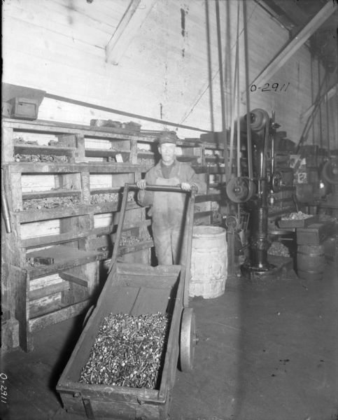 A man in overalls stands with a large cart filled with parts at International Harvester's Osborne Works (later known as Auburn Works). Behind him is a wall with wooden bins storing more parts. In the background on the floor on the right is a barrel and belt-driven machinery attached to the ceiling.