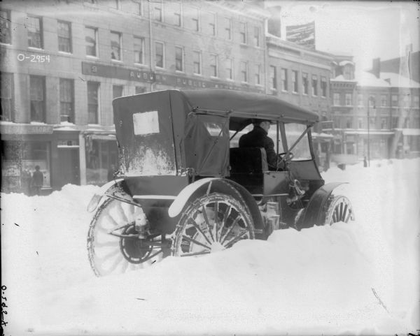 A man is sitting in the driver's seat of a vehicle that appears to be stopped in deep snow. There is a canvas covering over the top of the vehicle. In the background across the street are brick buildings and a few men in front of storefronts. The sign above the storefront reads: "Auburn Public Market?"