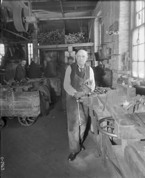 A man dressed in a vest, tie and a long apron stands near a vise at a work station at International Harvester's Osborne Works (later known as Auburn Works). Behind him are several other factory workers pushing carts filled with tools and parts. There is a coat and hat draped over one of the carts. Above the men is a loft filled with lumber, and belt-driven machinery attached to the ceiling.