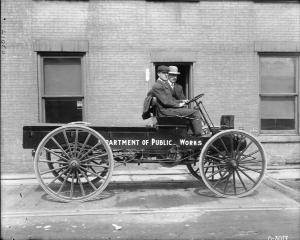 Two men are sitting in a truck with an empty flatbed and large wheels. The men are both wearing suits and hats. The sign on the side of the vehicle reads: "Department of Public Works." Behind the men is a large brick building.