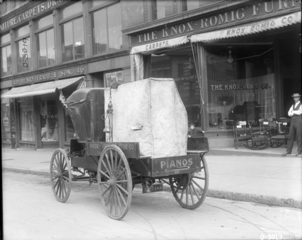 A delivery truck with the store name: "Knox-Romig Furniture Co." is parked in front of the Knox-Romig storefront. There is a covered piece of furniture, probably a piano, loaded in the truck. The word "Pianos" is painted on the back of the truck. The storefront awning advertises "Carpets" and features merchandise in the windows and in front of the store. A man dressed in dark pants, a vest, and a hat stands next to a group of chairs at the entrance to the store on the right. A young boy stands on the left side of the entrance.