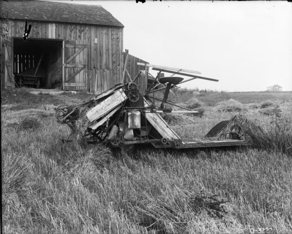 A grain binder (likely an Osborne) sitting in a field near a barn filled with piles of hay and a cart.