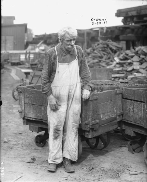 A man dressed in light overalls and a stripped button-up shirt is leaning against a cart in a factory yard at International Harvester's Osborne Works (later known as Auburn Works). The man has white hair and a large moustache. In the background is either rubble or building materials.