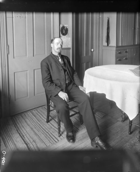 A man with a moustache dressed in a dark suit and necktie is sitting on a wooden chair next to a table covered with a light-colored tablecloth. The floor is covered in thin carpeting. The room has two doors and built-in cabinets. Behind the man a calendar is hanging on the wall.