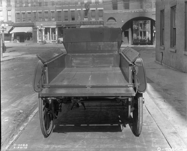 Rear view of the bed of an International(?) truck parked on a city street. In front of the truck is a block filled with storefronts. Streetcar tracks run along the street and through an arch set in the block of buildings in the background. This photograph may have been taken in Auburn, New York.