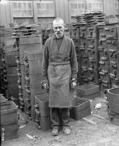 A man dressed in work clothes and an apron stands in a factory yard at International Harvester's Osborne Works (later known as Auburn Works). Behind the man are stacks of what appear to be building materials or machine parts, and the factory wall beyond. Foundry area.