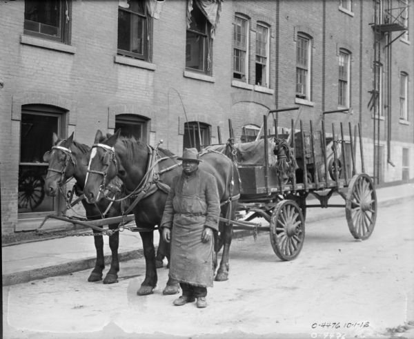 An African American man dressed in work clothes, a long apron, and a hat is standing next to a cart drawn by two horses. The cart is loaded with goods and rope. Behind the cart is a large brick building, probably a factory building at International Harvester's Osborne Works (later known as Auburn Works).