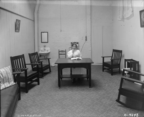 A woman, probably a factory worker at International Harvester's Osborne Twine Mill, is sitting at a wooden desk reading a book. She is in a narrow room with a tall ceiling. The room is furnished with several chairs and a couch. Behind the woman is a sink with a mirror hanging above it, a chamber pot, and a telephone. On either side of the room paintings are hanging on the walls, and a window at the top of the room is curtained. The room is likely a factory break room.