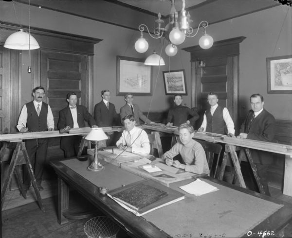 A group of nine men are gathered in a room, possibly a classroom. Seven  of the men are standing at a makeshift table on sawhorses on the edges of the room. The other two men are seated at a large table with a blotter, lamp, and drawing materials. Below the table is a wastebasket. There are three large light fixtures hanging from the ceiling, and three framed prints on the walls. The room may be part of International Harvester's Osborne Works (later known as Auburn Works) factory.