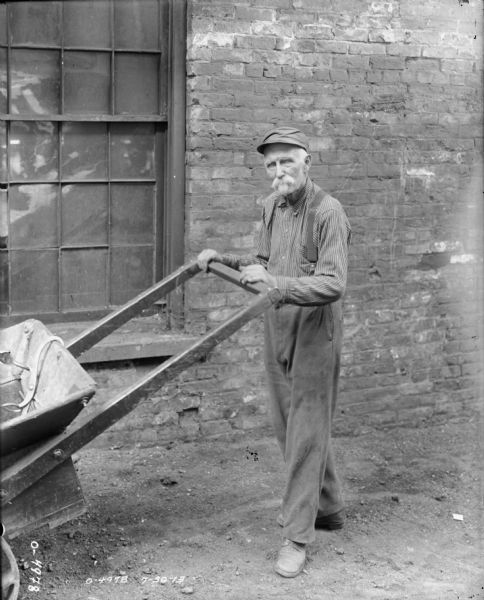 An elderly factory worker dressed in pants, a striped shirt, suspenders and a hat is posing with a large push cart at International Harvester's Osborne Works (later known as Auburn Works). Behind the man is a brick building, probably part of the factory.
