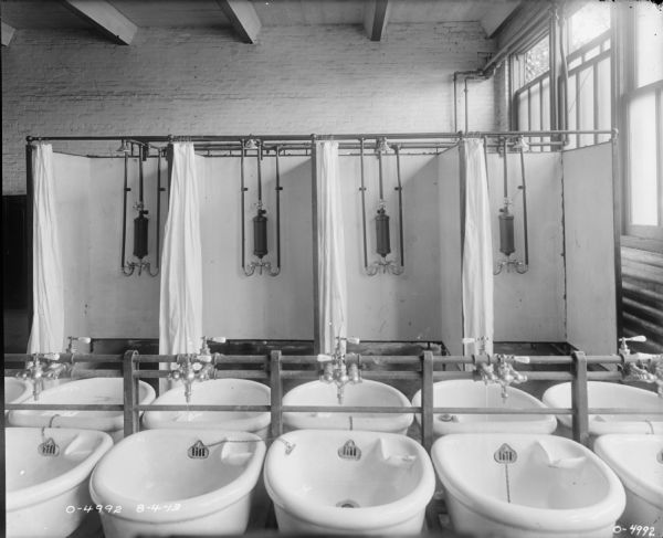 A large factory washroom is equipped with a double row of sinks, and along a wall, four showers at International Harvester's Osborne Works (later known as Auburn Works). The room is large with a tall ceiling and large windows.