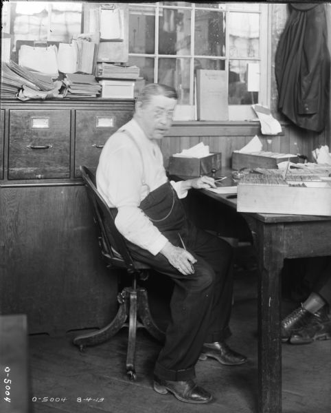 A man wearing glasses is seated at a cluttered wooden desk at International Harvester's Osborne Works (later known as Auburn Works). Behind him are file drawers and stacks of papers. An unseen person is on the other side of the desk, as a second pair of feet can be seen underneath the table. A suit jacket is hanging on the wall near a window.