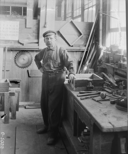 A factory worker in work clothes and a hat stands at a work station at International Harvester's Osborne Works (later known as Auburn Works). The wooden table is scattered with tools, and on the wall behind him hangs a saw blade and calendar. To the right is a window with jars and bottles on the windowsill.