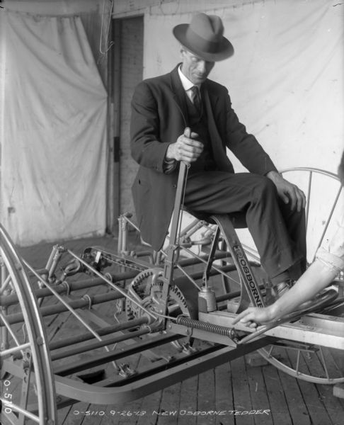 A man dressed in a suit and hat is sitting on a New Osborne hay tedder at International Harvester's Osborne Works (later known as Auburn Works). On the right the arm of another man is pointing at a spring on the tedder. Two light-colored sheets are hanging as a makeshift background. The tedder has an "Osborne" decal on it.