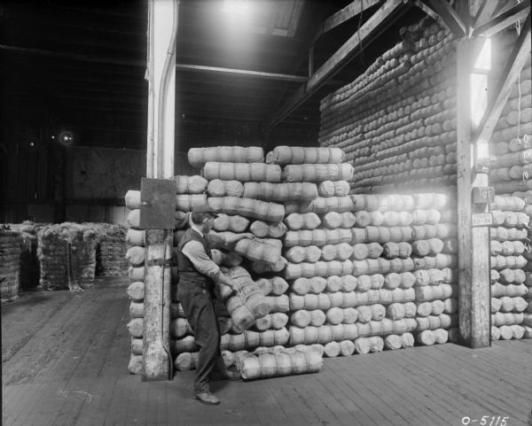 A factory worker stacks wrapped and tied bundles of twine into a large pile in the Twine Mill at Osborne Works (later known as Auburn Works). Behind the man are tied up piles of henequen or sisal fiber. There is a skylight in the high ceiling.