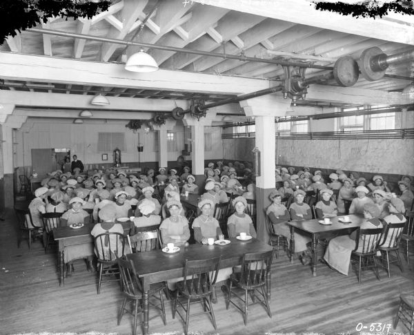 Elevated view of a large group of female factory workers sitting at tables in a cafeteria at International Harvester's Osborne Works. The women likely worked in the Twine Mill. The women are dressed in work clothes and puffy hats. The room has several rows of long wooden tables and chairs, and the tables are set with tableware. On the back wall is a pail, a large metal urn, and a Victrola. Two or three people, perhaps kitchen staff, are standing at a doorway in the back. The room has a high ceiling with exposed rafters and pipes, electric lights hang from the ceiling, and there are narrow windows at the top of the room.