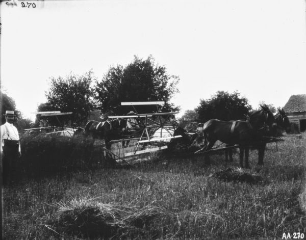 A man and a young boy are each driving a horse-drawn binder in a field. A man wearing a hat and suspenders is standing in the foreground on the left. On the far right is a building.
