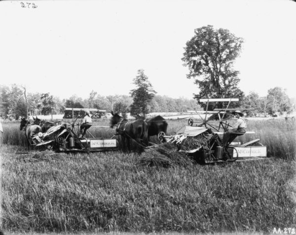 View of two young men with two horse-drawn binders in a field. There is a wooden fence and trees behind them.