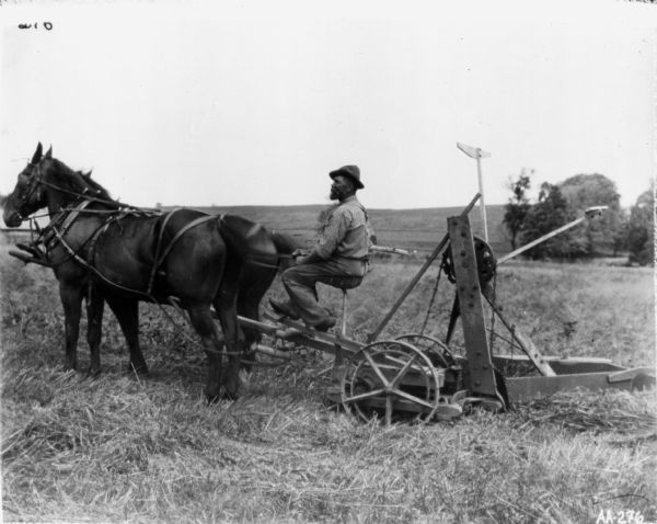 Bearded man operating a reaper drawn by two brown horses. The reaper appears to be an early model or may be a replica of the first reaping machine.  
