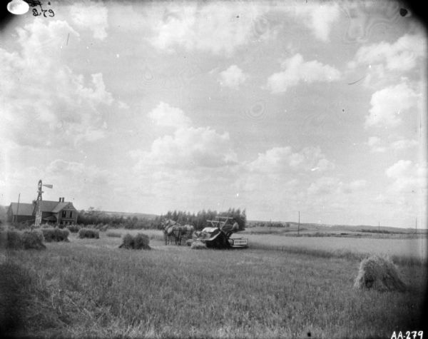 View across field towards a man using a horse-drawn binder. There is a farmhouse, farm buildings and a windmill in the background on the left.