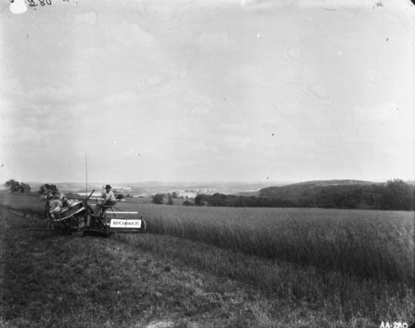 Rear view of a man using a horse-drawn binder in a field. In the background are hills and a valley.