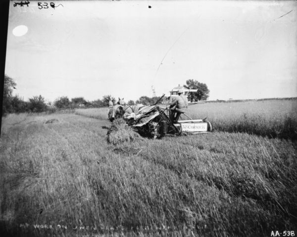 Three-quarter view from rear of a man using a horse-drawn McCormick binder on the Gary Owen farm. A farmhouse and windmill are in the far background.
