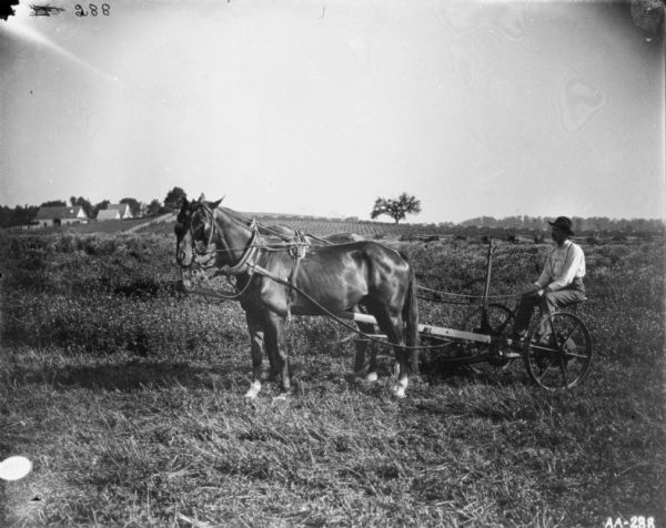 Man with horse-drawn mower in field. Farm buildings are in the background on the left.
