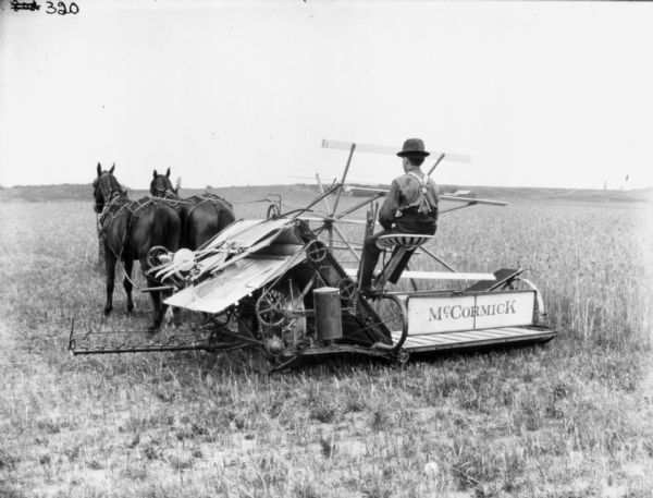 Rear view of young man using a McCormick horse-drawn binder in a field. There are farm buildings in the far background.