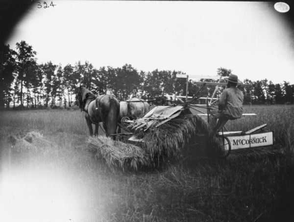 Three-quarter view from left of a man using a McCormick horse-drawn binder in a field. Sheaves of wheat are lyying on the ground. In the background is a row of trees.