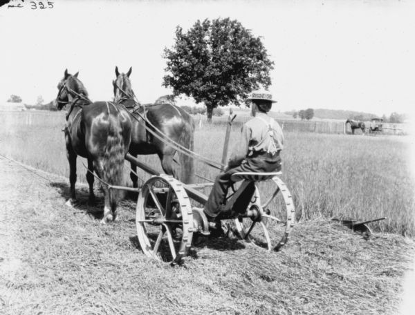 Three-quarter view from rear left side of a man using a horse-drawn mower in a field. A horse-drawn buggy is in the background on the right.