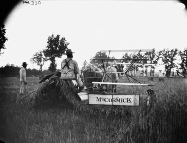 View from rear of a man using a horse-drawn binder in a field. Another man is standing on the left nearby.