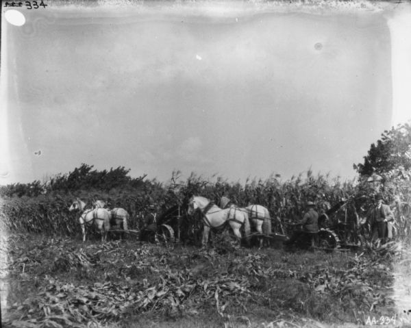 Two men using horse-drawn corn binders in a field. A man wearing a suit and hat is standing on the right.